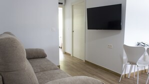 A living room with a beige sofa, a flat-screen TV mounted on the wall, and a white chair.