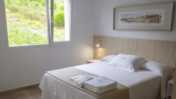 A neatly made bed with white linens and towels, a bedside table with a lamp, and a window with a view of greenery.