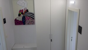 Quarto de hotel com um guarda-roupa branco, um banco e uma obra de arte colorida na parede.