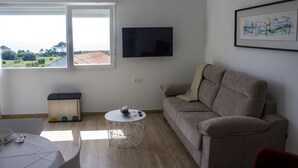 A living room with a sofa, a round coffee table, a TV mounted on the wall, and a window with a view of the outdoors.