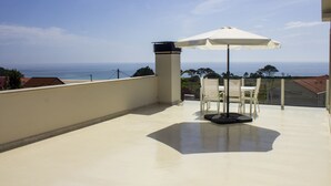 A balcony with a white umbrella, white chairs, and a view of the sea.