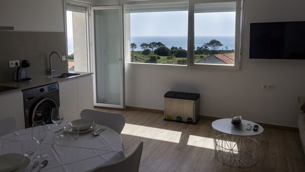 A modern kitchen with a washing machine, a dining area with a table set for four, and a large window offering a view of the sea.