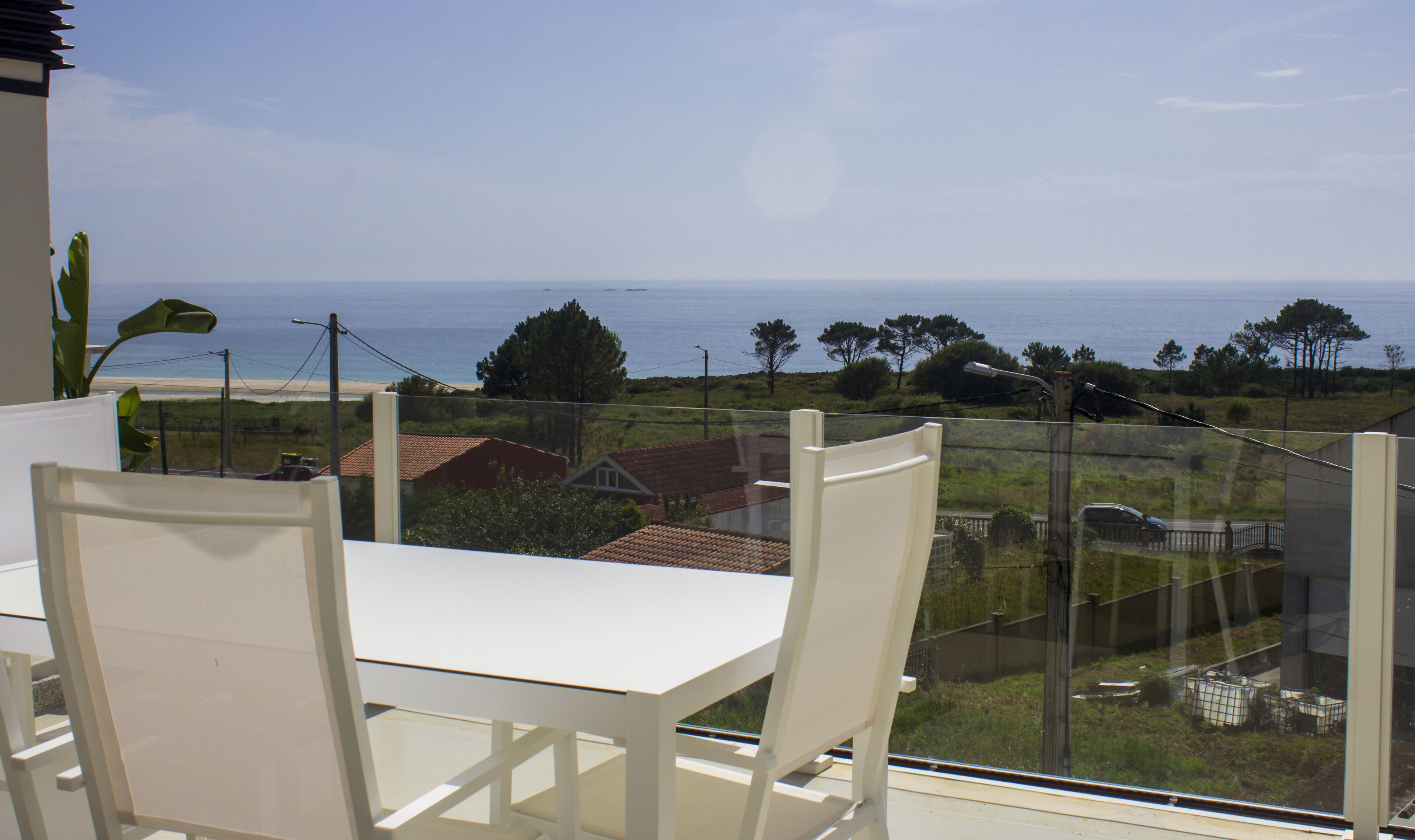 A balcony with a white table and chairs, overlooking a coastal view with houses and greenery.