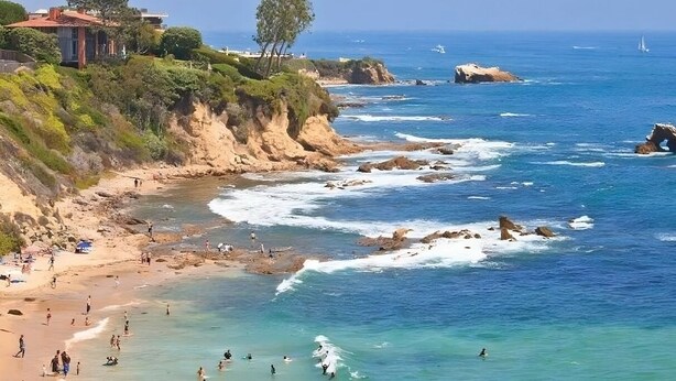 Une scène de plage côtière avec des falaises rocheuses, une eau bleue et limpide, et quelques personnes qui profitent de la plage.