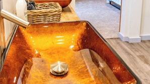 Ein Badezimmer mit einem großen, rechteckigen, bernsteinfarbenen Waschbecken, Holzboden, weißer Wand mit einem Gemälde und einer Holztür.