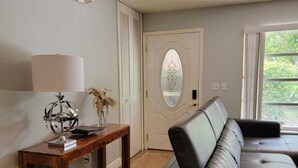 A living room with a leather sofa, a wooden side table with a globe and lamp, a door with a glass panel, and a window with a view of greenery. - Spacious 4-bedroom house in tranquil Vero Beach with AC, WiFi (Vero Beach)