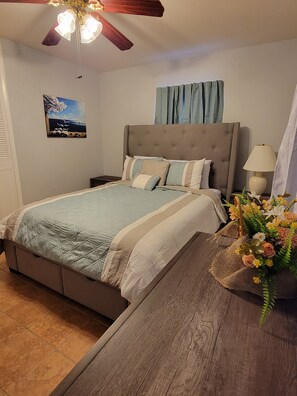 A bedroom with a bed, a nightstand, a lamp, a ceiling fan, a painting, and a window with curtains.