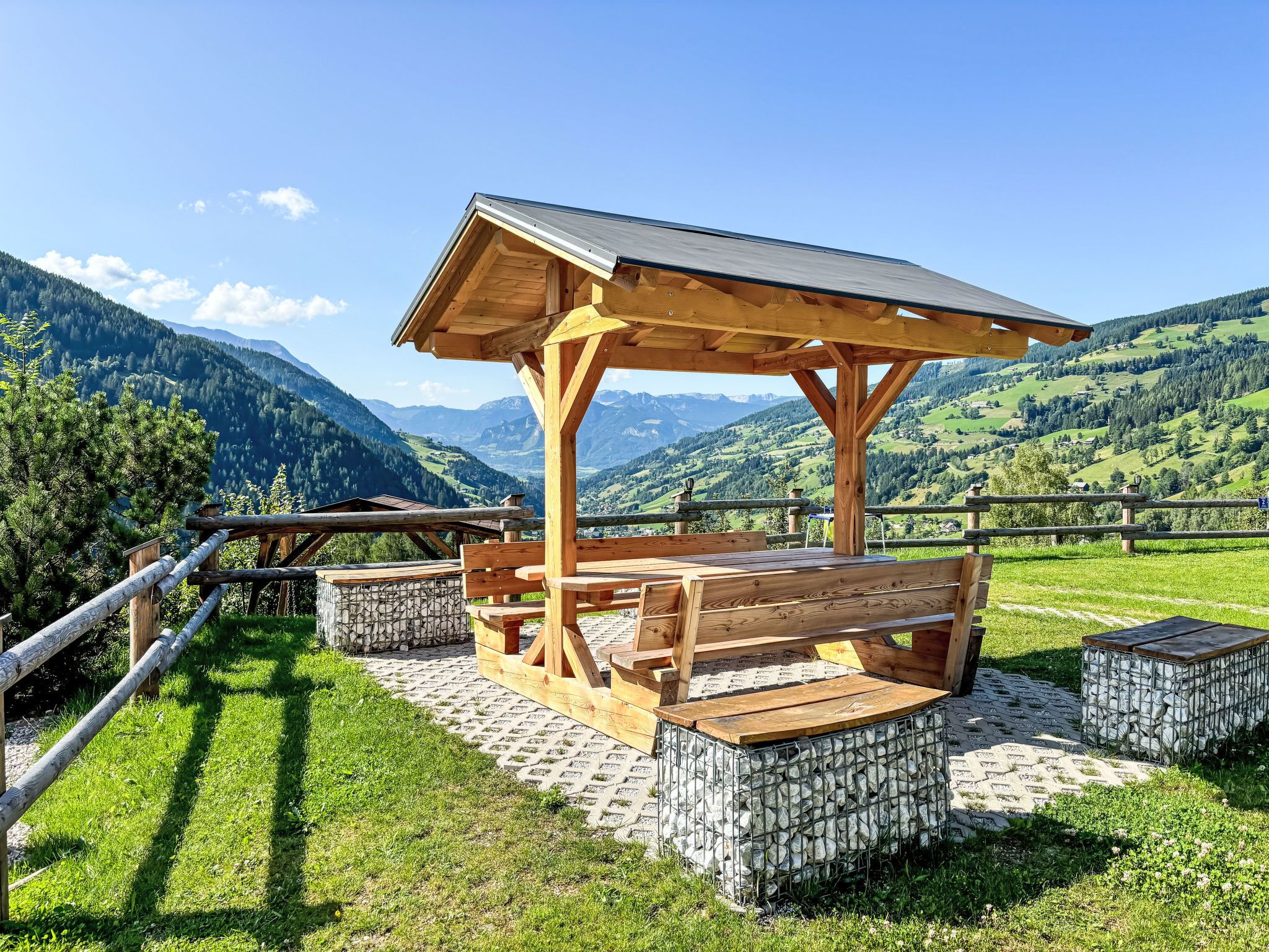 Um gazebo de madeira com bancos e uma mesa de pedra, situado numa área pavimentada com vista para a montanha.