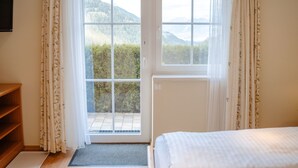 Een kamer met een bed, een houten plank, een raam met uitzicht op bergen en een deur met gordijnen.