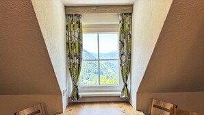 Una habitación con una mesa de comedor de madera, un ventanal con vistas a una cadena montañosa boscosa y una cortina con estampado de hojas.