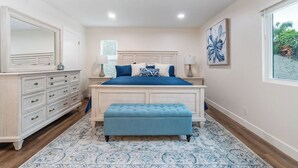 Un dormitorio con cama, mesitas de noche, cómoda, banco y un ventanal grande.