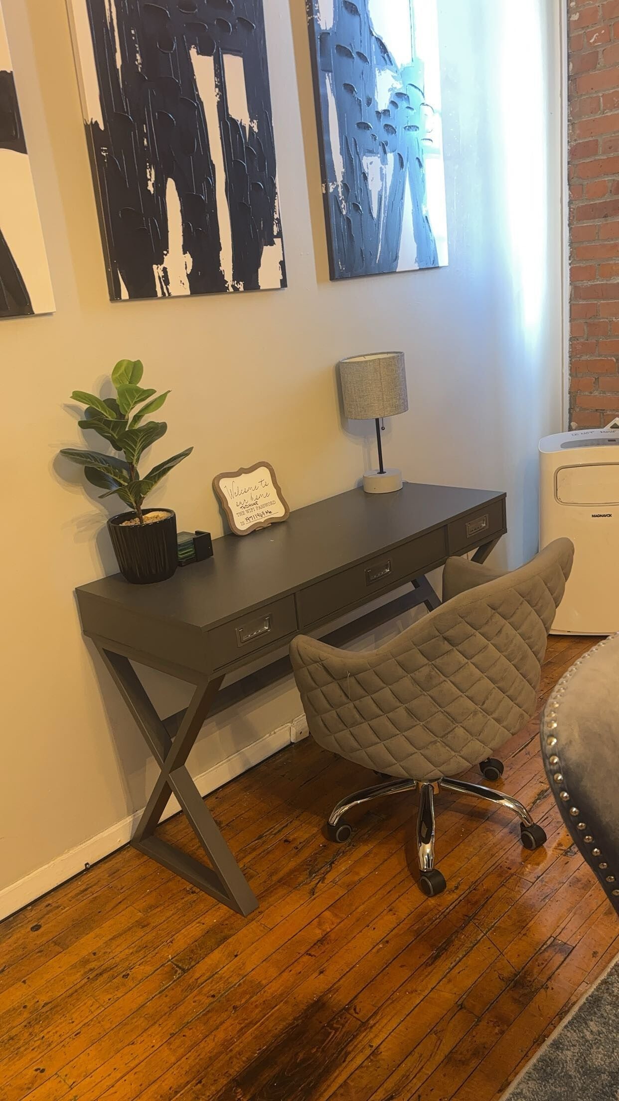Un espace de bureau moderne avec un bureau gris foncé, un fauteuil de bureau confortable, une plante en pot et une œuvre d’art abstraite accrochée au mur.