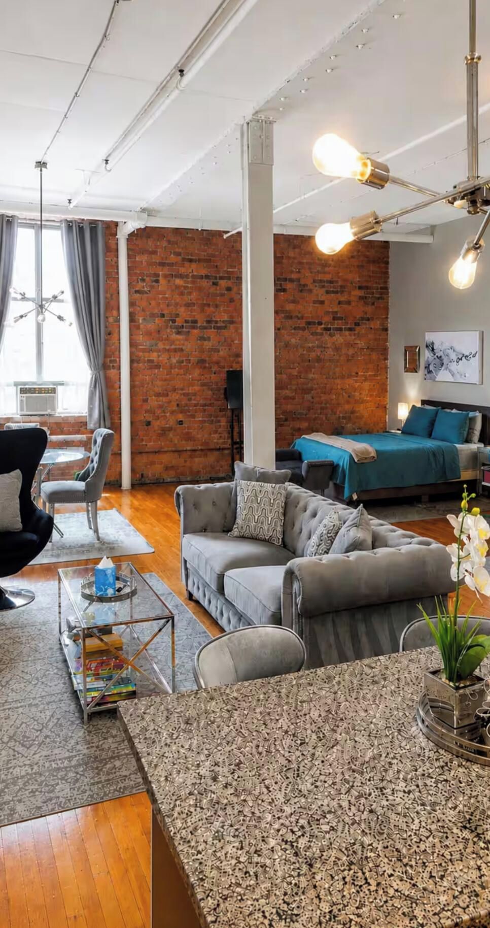 A modern living room with a brick wall, a sofa set, a coffee table, and a bed with a blue bedspread.