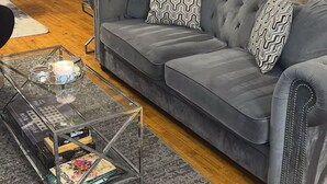 Un salon avec un canapé gris, des coussins à motifs, une table basse en verre sur laquelle sont posés des magazines et un mur en briques.