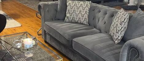 Ein Wohnzimmer mit grauem Sofa, gemusterten Kissen, einem Glastisch mit Zeitschriften und einer Ziegelwand.