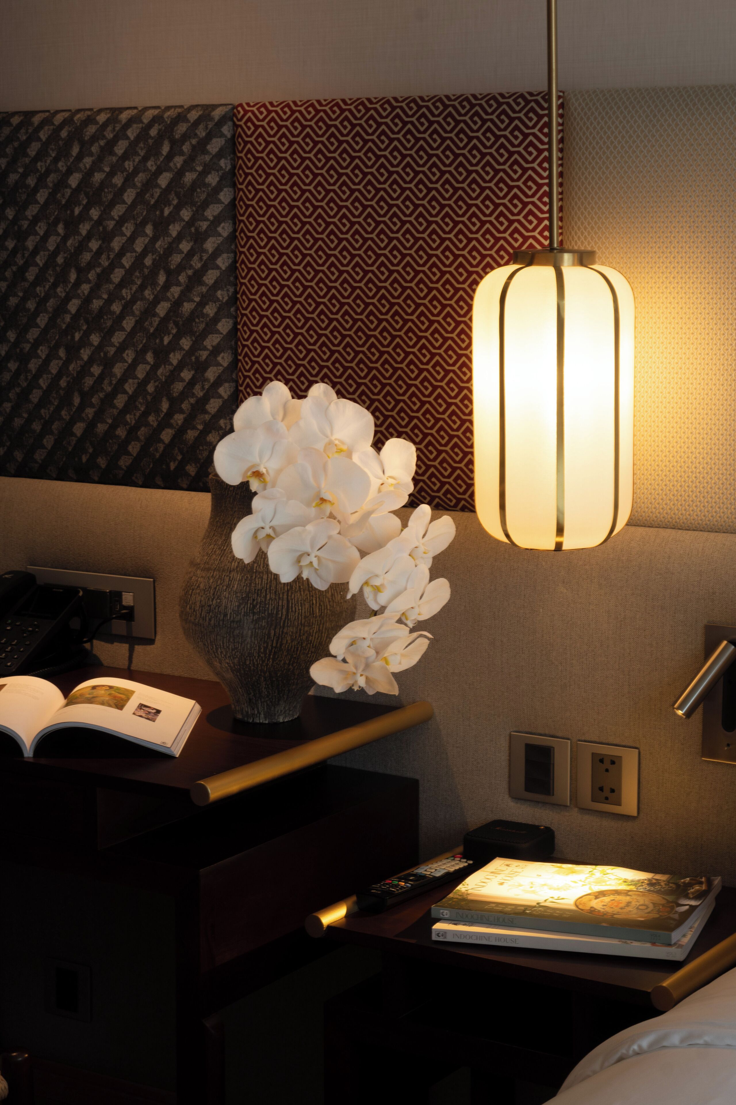 Een hotelkamer met nachtkastje, een vaas witte orchideeën, een boek, een afstandsbediening en een lamp.