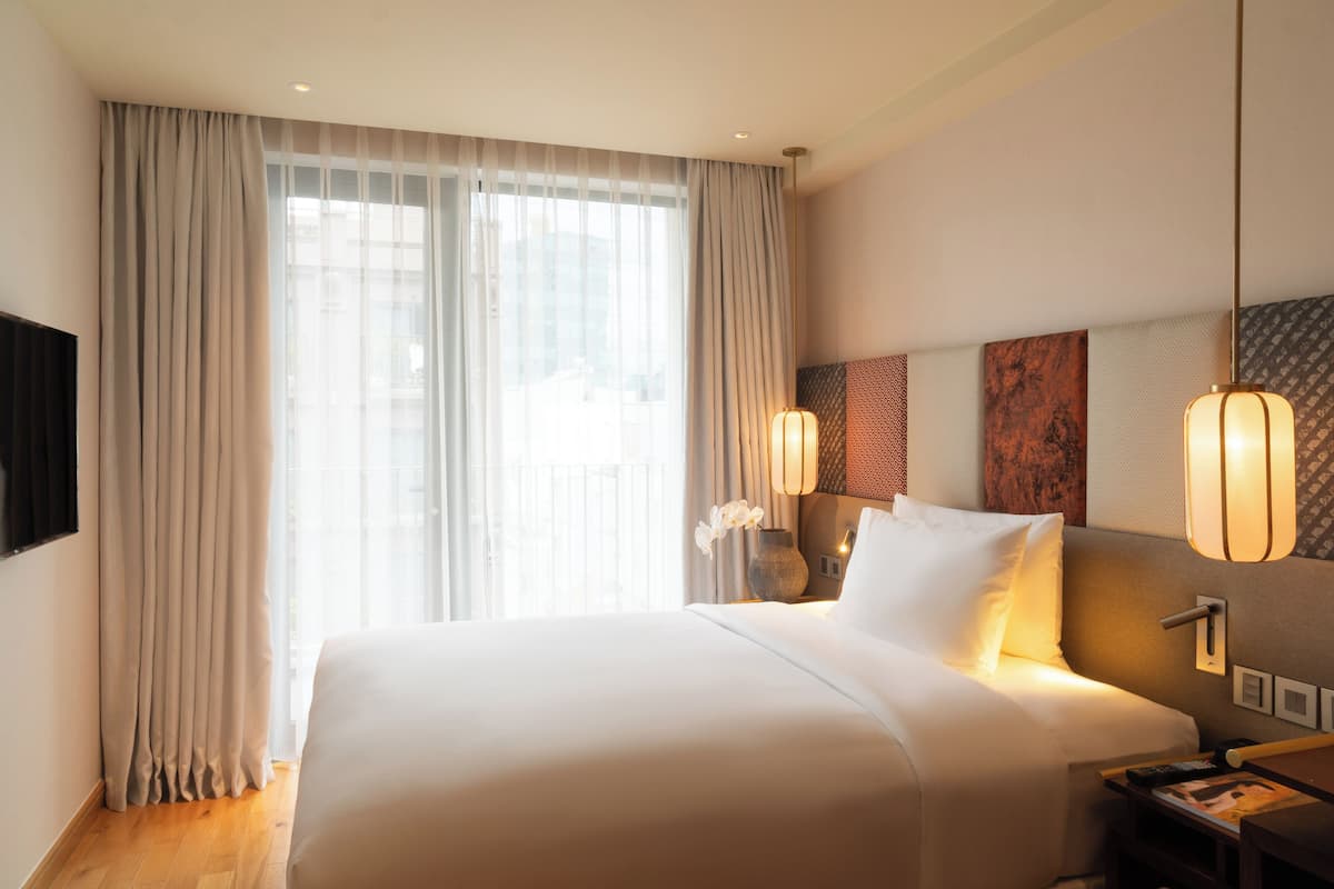 Ein Hotelzimmer mit einem großen Bett, einem an der Wand montierten Fernseher, einem Nachttisch mit Lampe und Blick auf die Stadt durch das Fenster.