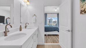 A modern bathroom with a large sink, two faucets, and a mirror. There is a door leading to a bedroom with a bed and a view of buildings outside. - 3 Stories w/86" TV | Keurig | Pro Cleaned | Garage (Seaside Heights)