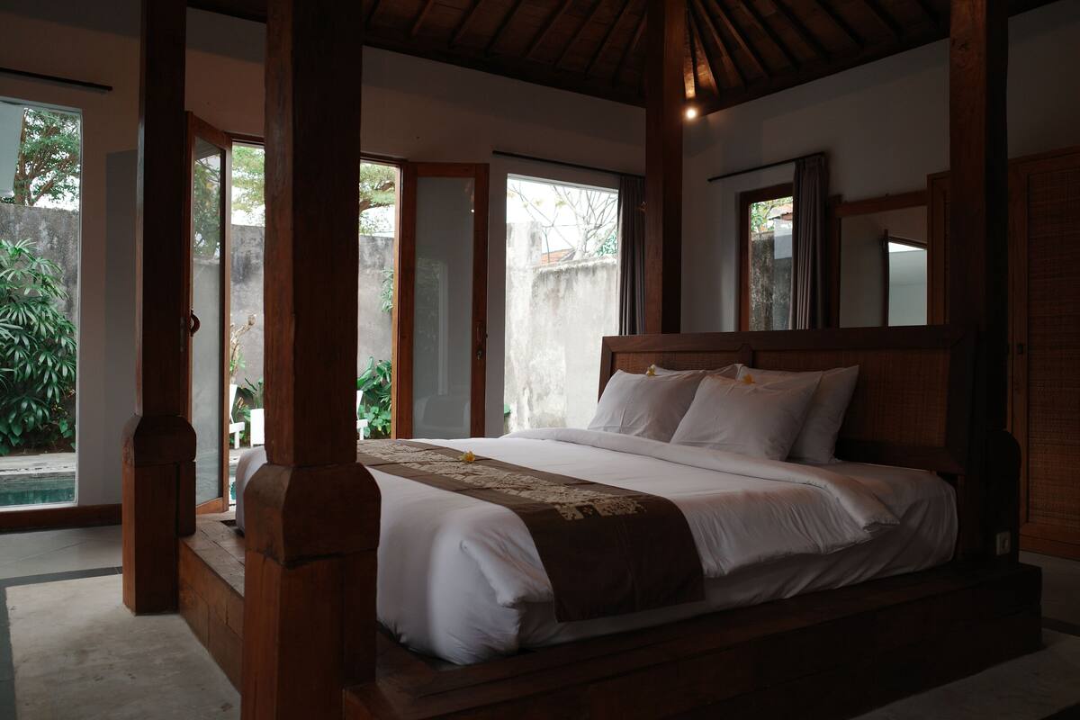 Una cama grande con cabecera de madera, una mesita de noche y vista a una piscina.