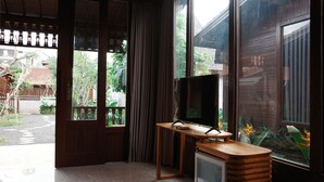 A room with a television on a wooden stand, a small refrigerator, and large windows with curtains. - The Riang Villa (Kerobokan)