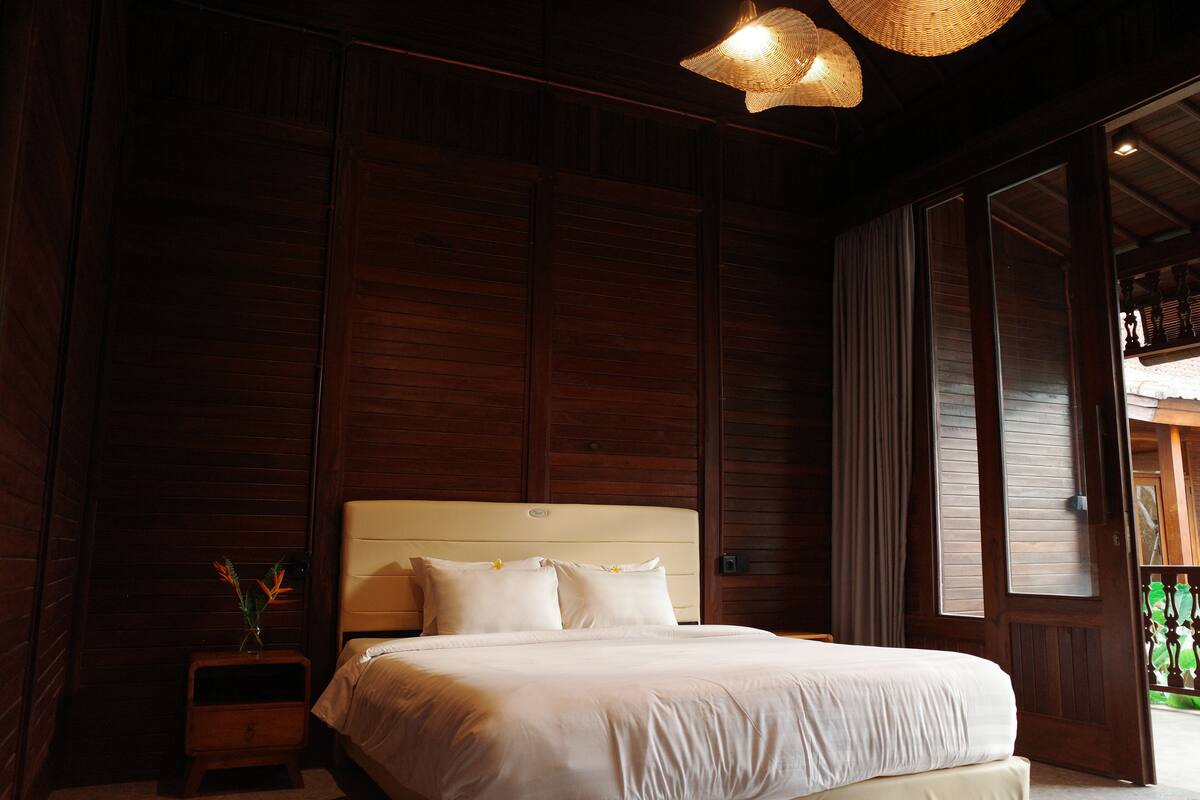 Un dormitorio con una cama grande, paneles de madera, una mesita de noche con un florero y una lámpara colgante.