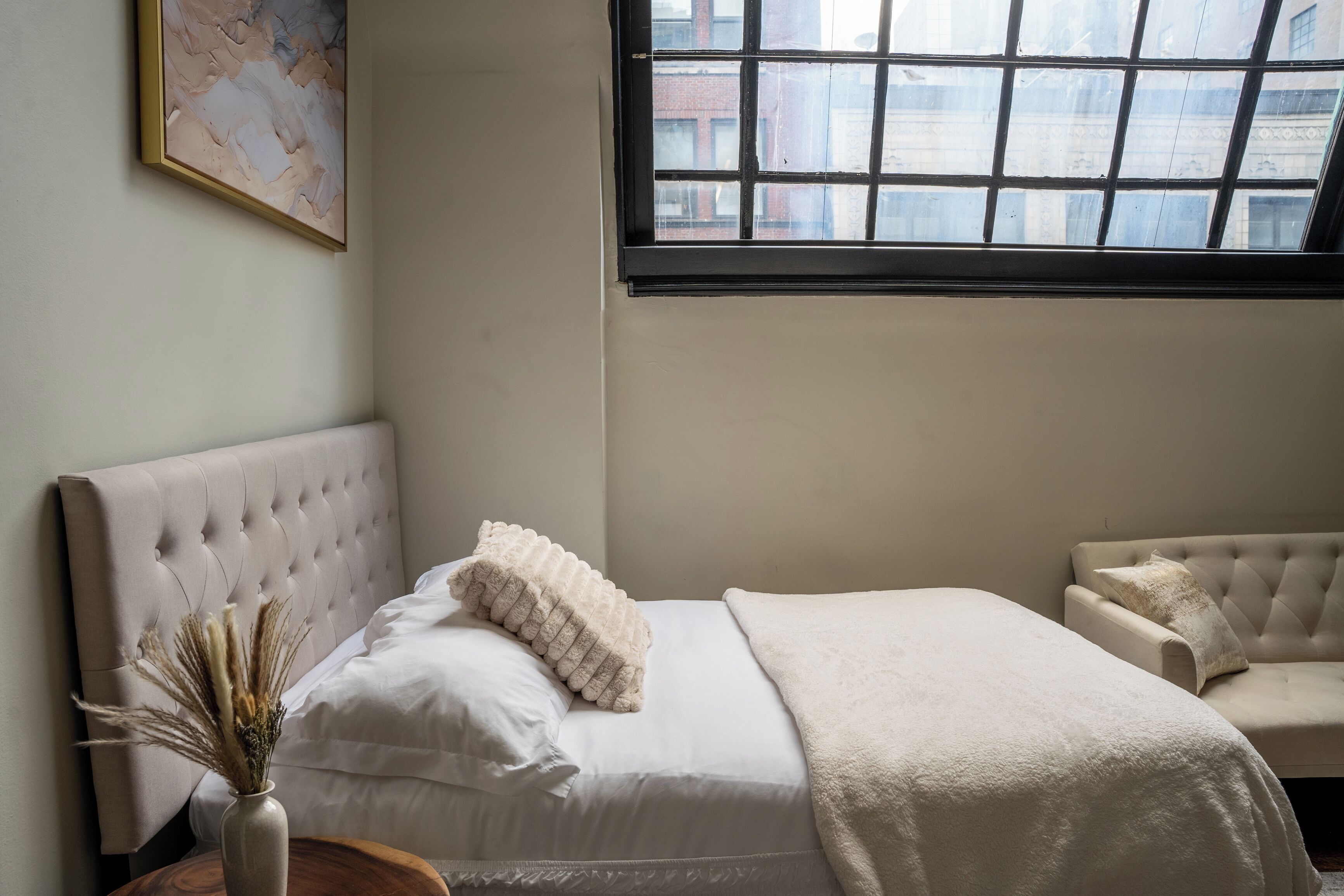 A bedroom with a large window, a tufted headboard, a white bed, and a beige armchair.