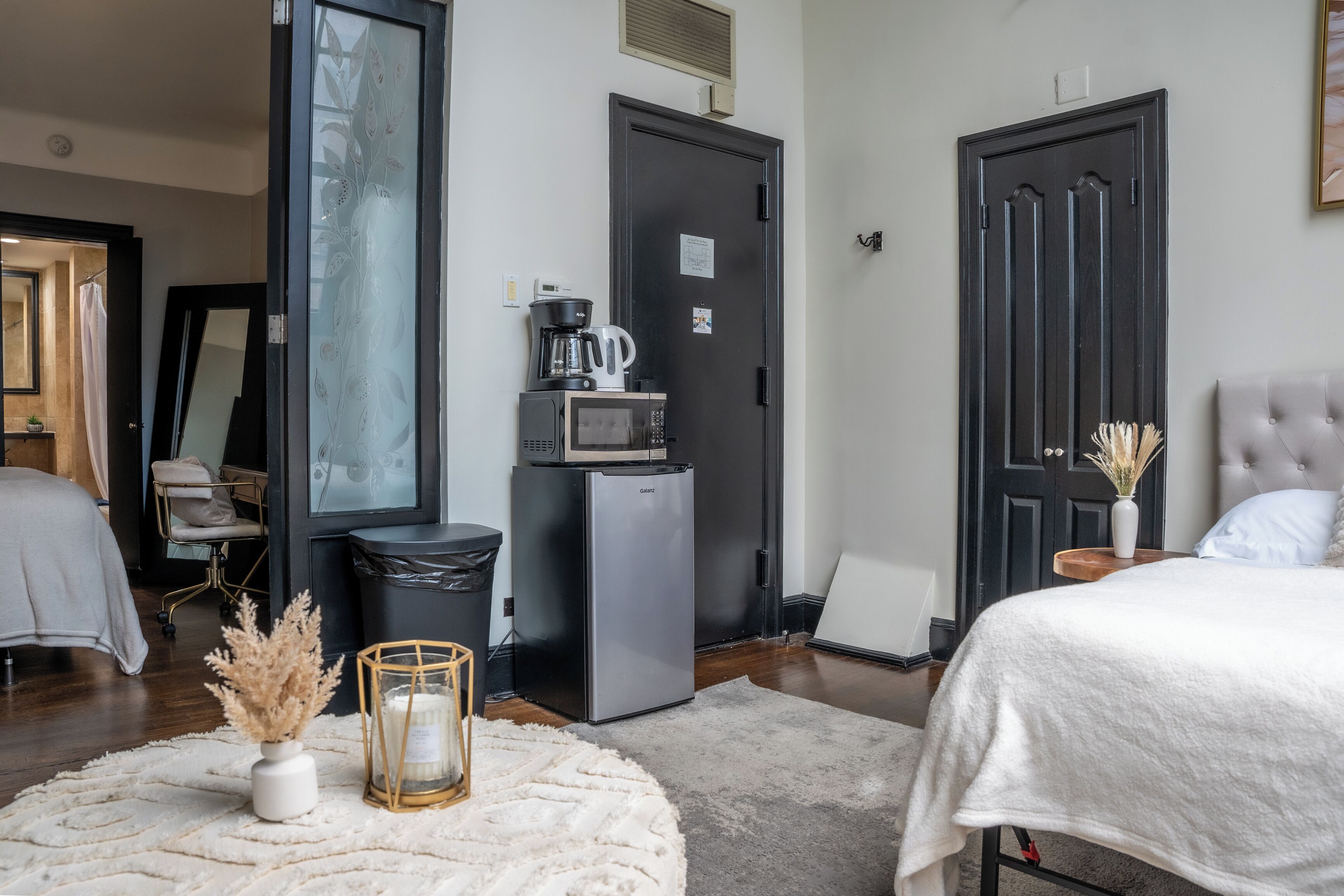 A modern hotel room with a bed, a small kitchenette, a trash bin, and a door with a frosted glass panel.