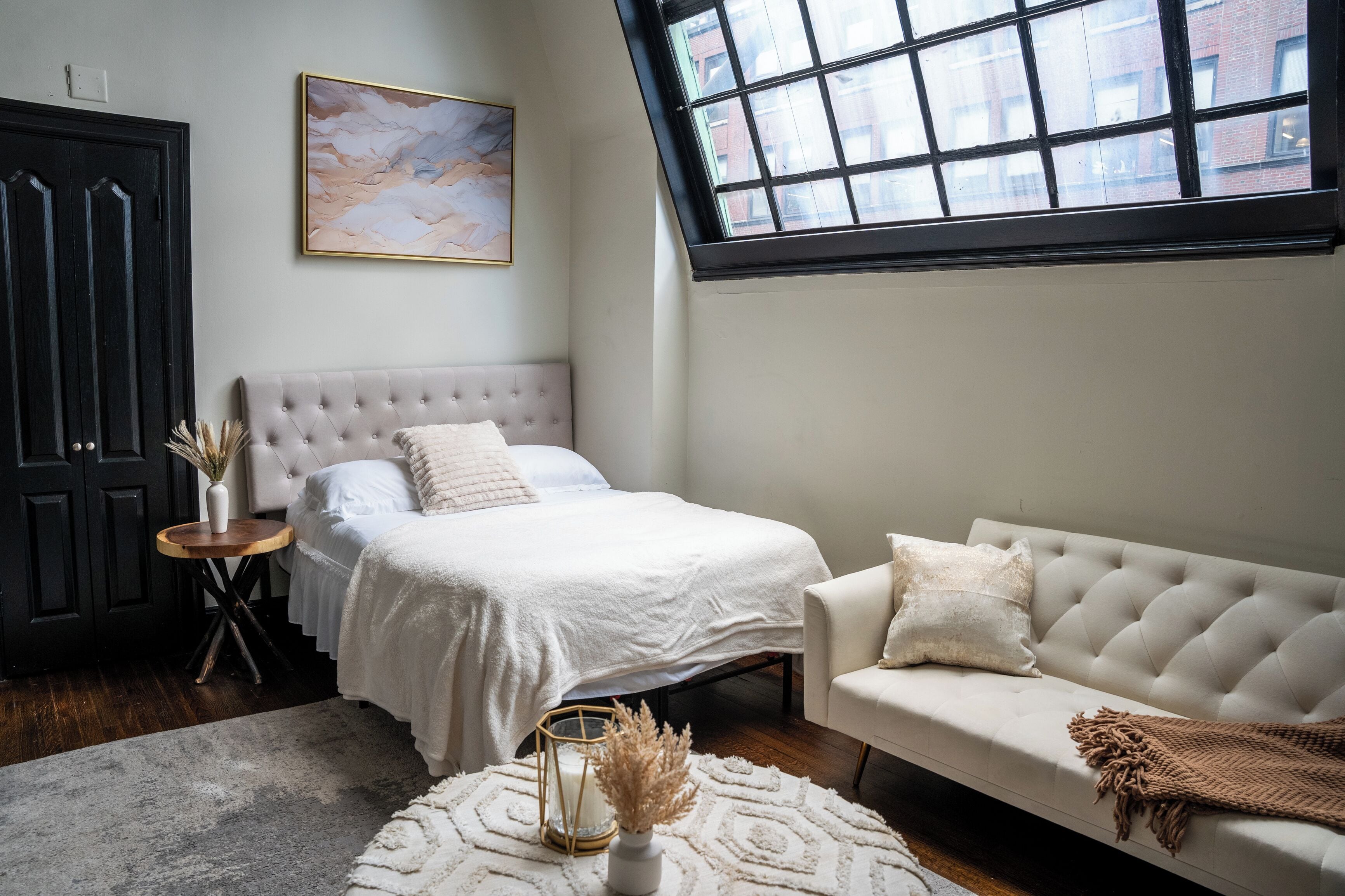 A bedroom with a bed, a white sofa, a window, and a painting on the wall.