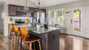 A modern kitchen with a central island, bar stools, stainless steel appliances, and a view through a door.