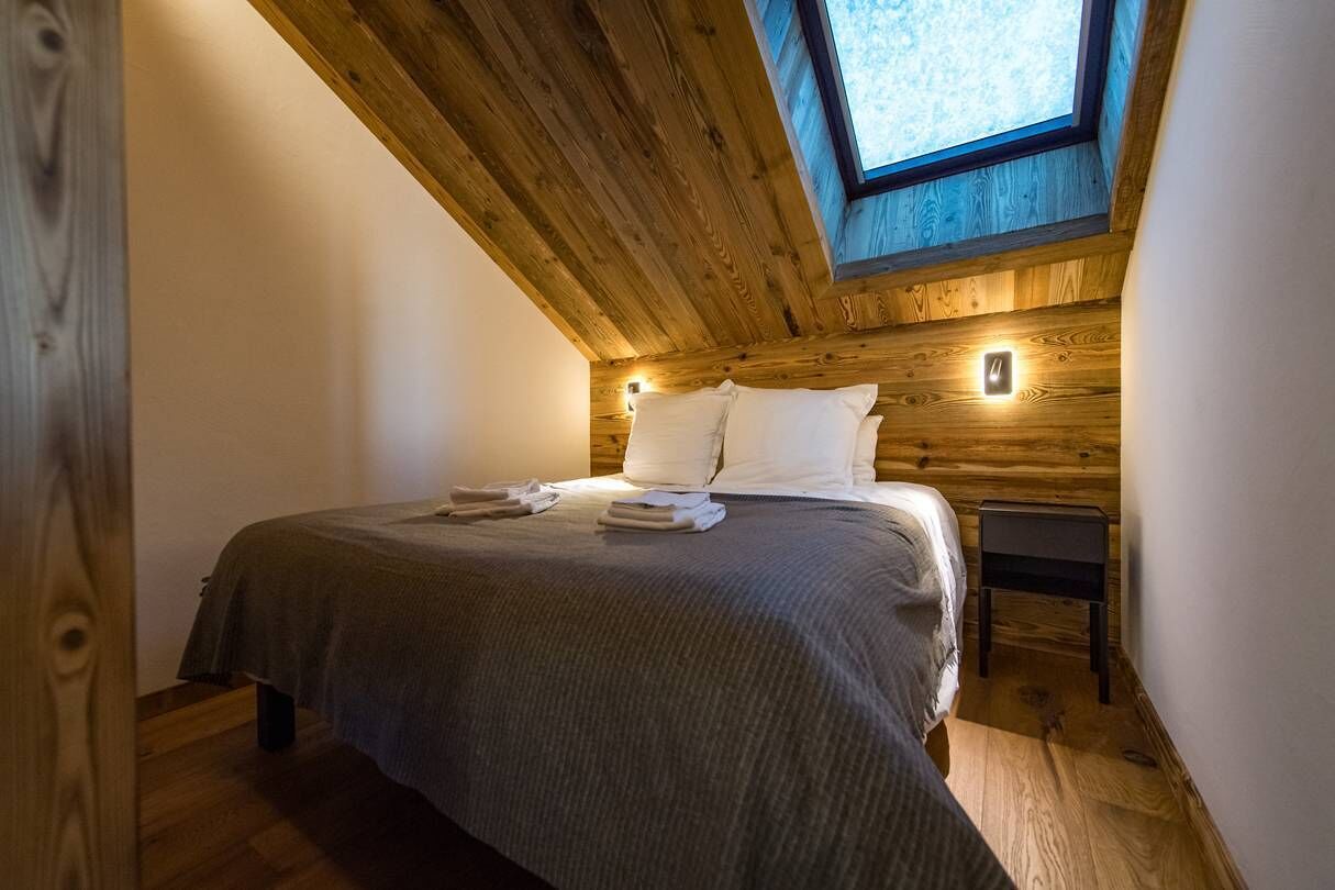 Ein Schlafzimmer mit Holzboden, einem Oberlicht, einem Bett mit Kissen und Decke sowie einem Nachttisch mit Lampe.