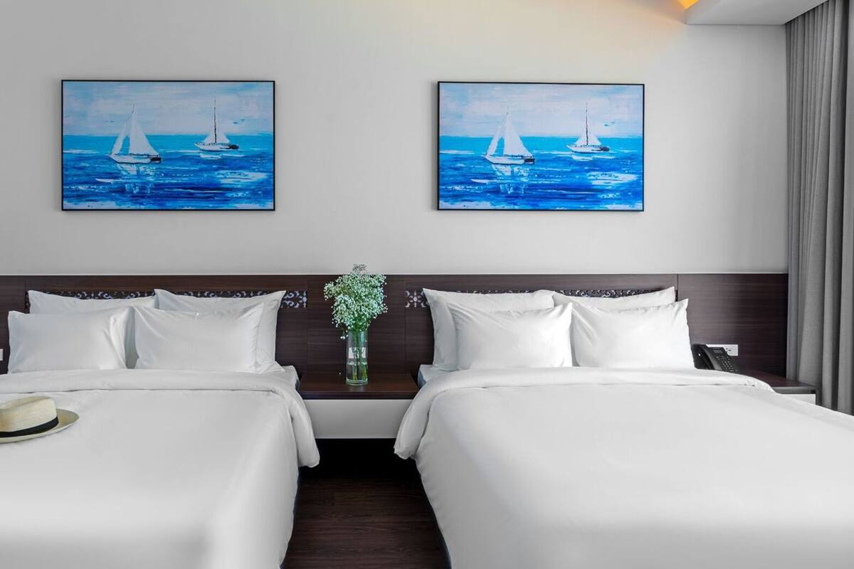 A hotel room with two beds, a nightstand, a vase with flowers, and two paintings of sailboats on the wall.