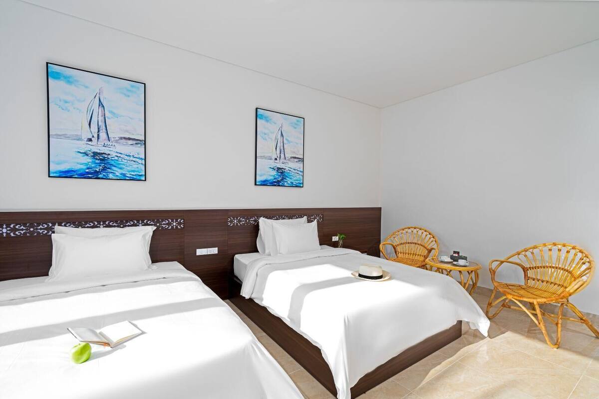 A hotel room with two beds, a wooden headboard, white bedding, two framed paintings of sailboats, a wicker chair, a small round table, and a green apple on the bed.