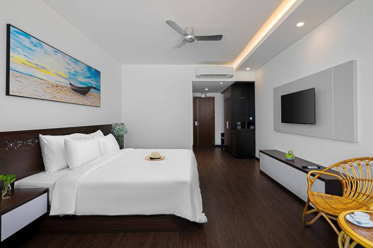 A modern hotel room with a bed, a TV, a dining area, and a hallway leading to a bathroom.