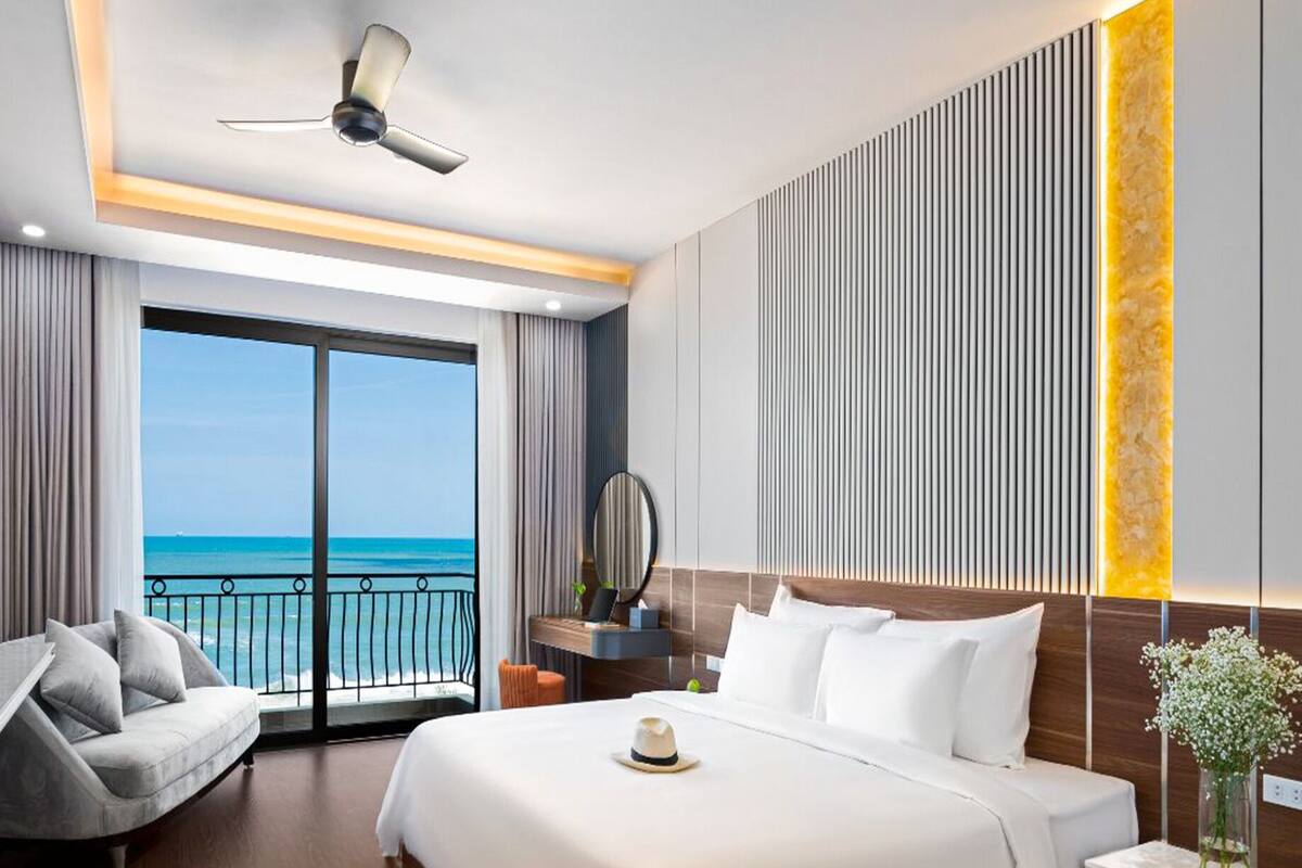 A hotel room with a bed, a TV, a ceiling fan, a desk, and a view of the ocean.
