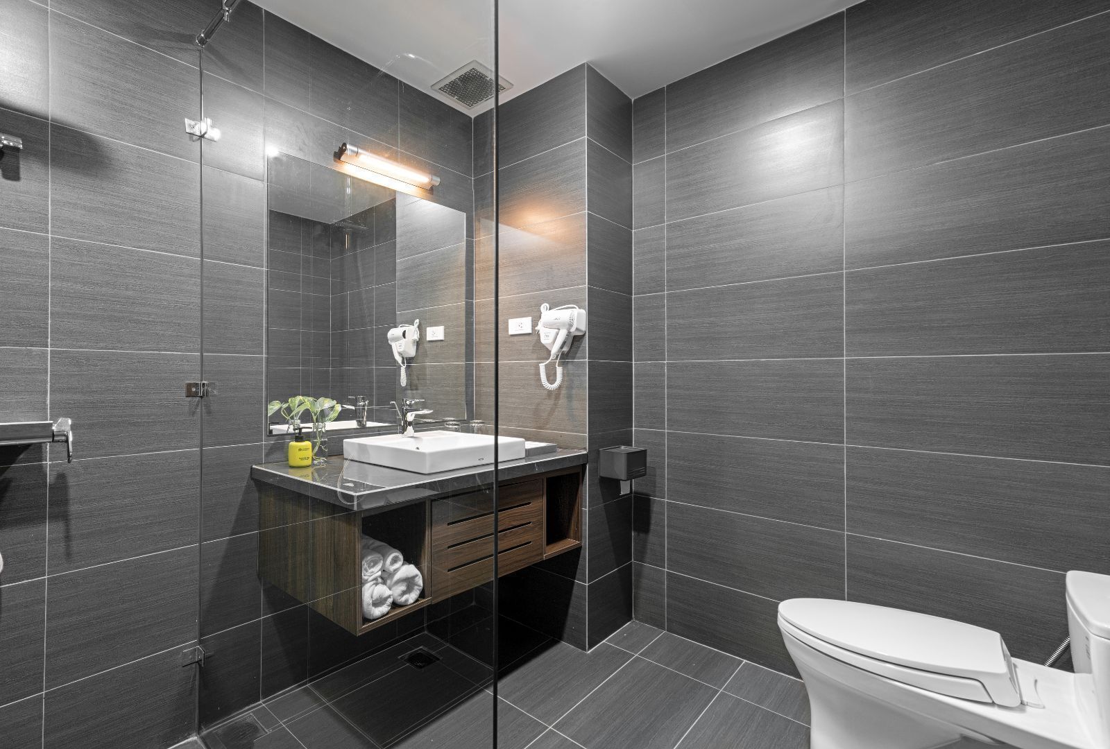 Un baño moderno con baldosas oscuras, ducha con mampara de vidrio, lavamanos blanco y un inodoro.