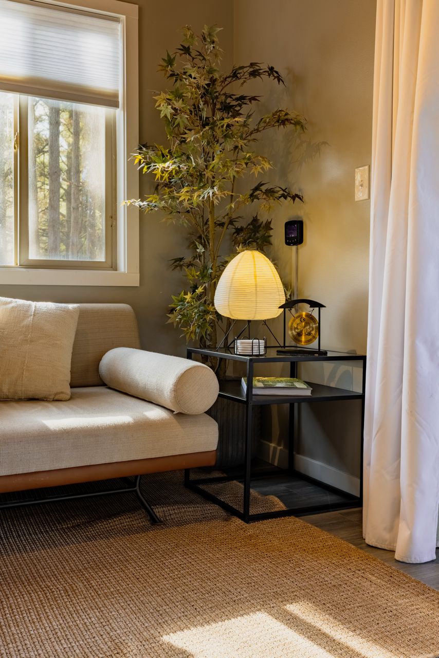 Une pièce avec un canapé, une lampe, une plante et une fenêtre avec des stores.