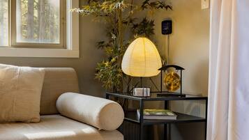 Une pièce avec un canapé, une lampe, une plante et une fenêtre avec des stores.