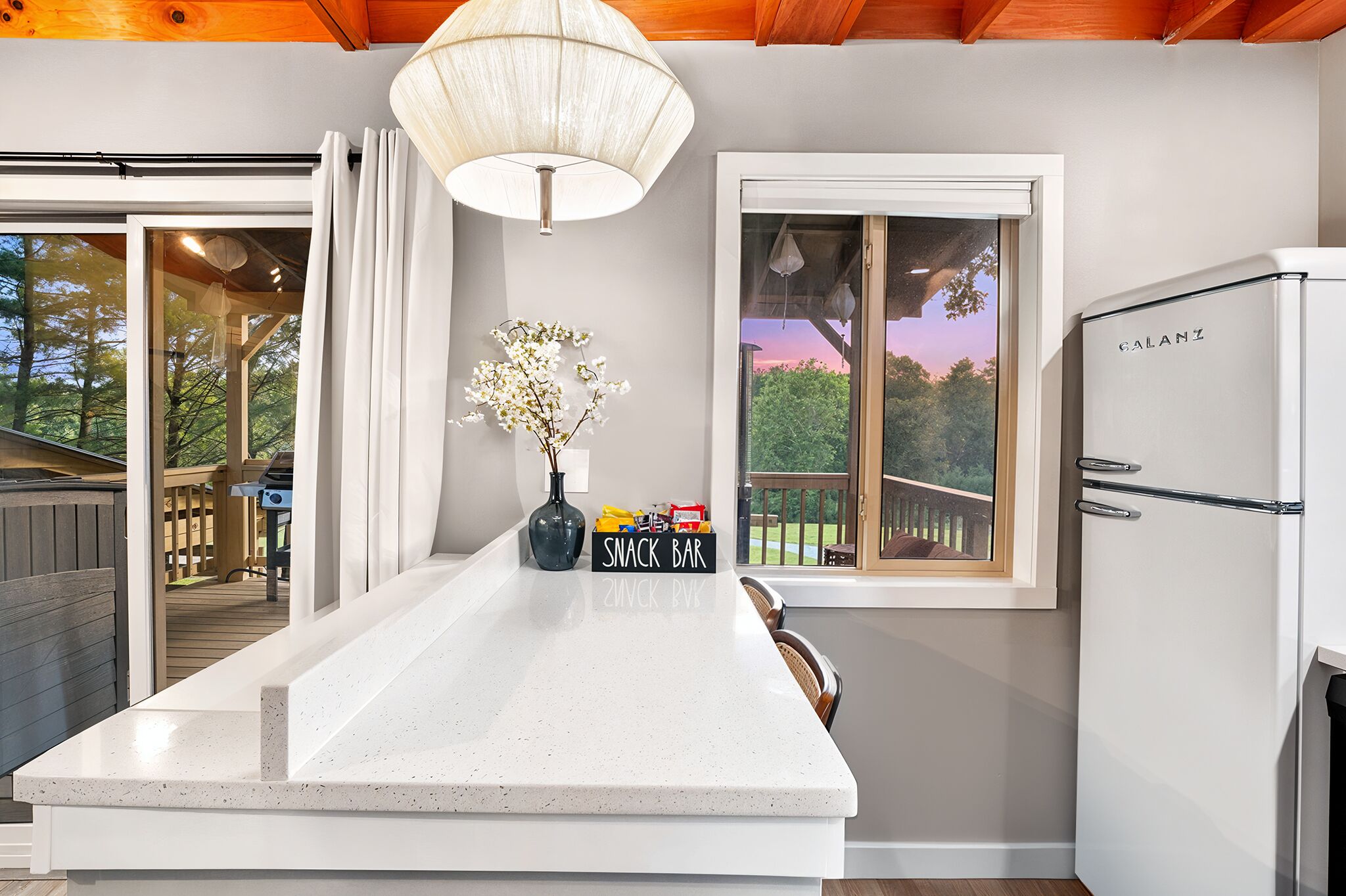 A kitchen with a white countertop, a 'SNACK BAR' sign, a refrigerator, and a window with a view of trees and a deck.