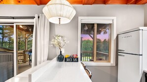 Una cocina con encimera blanca, un letrero que dice 'SNACK BAR', un refrigerador y una ventana con vista a árboles y una terraza.