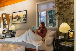 A person reading a book on a sofa in a room with a painting, a window with a view of trees, and a lamp on a side table.