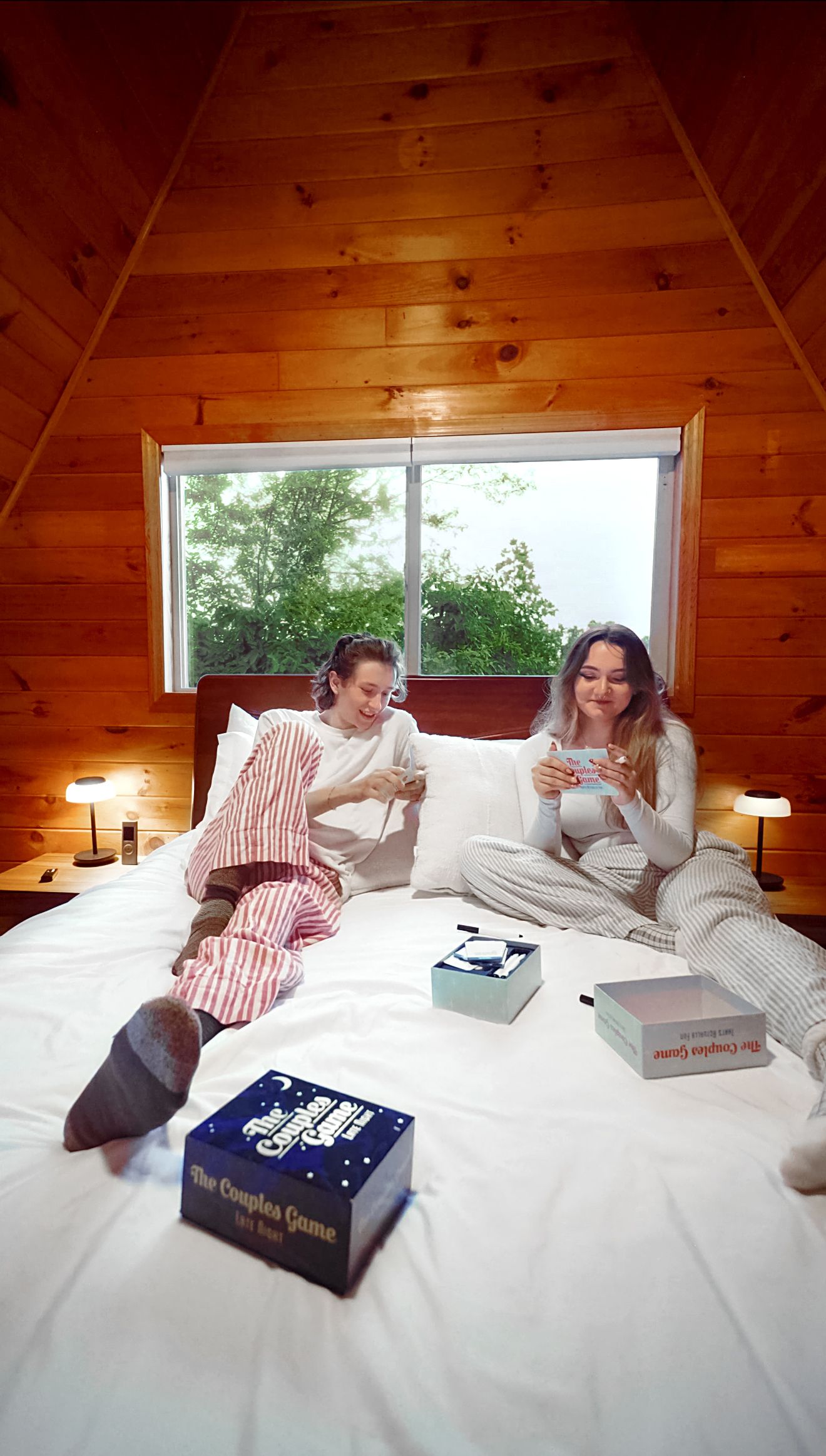 Une chambre avec un plafond en pente, une grande fenêtre, un lit avec des oreillers, et deux personnes qui lisent des livres.