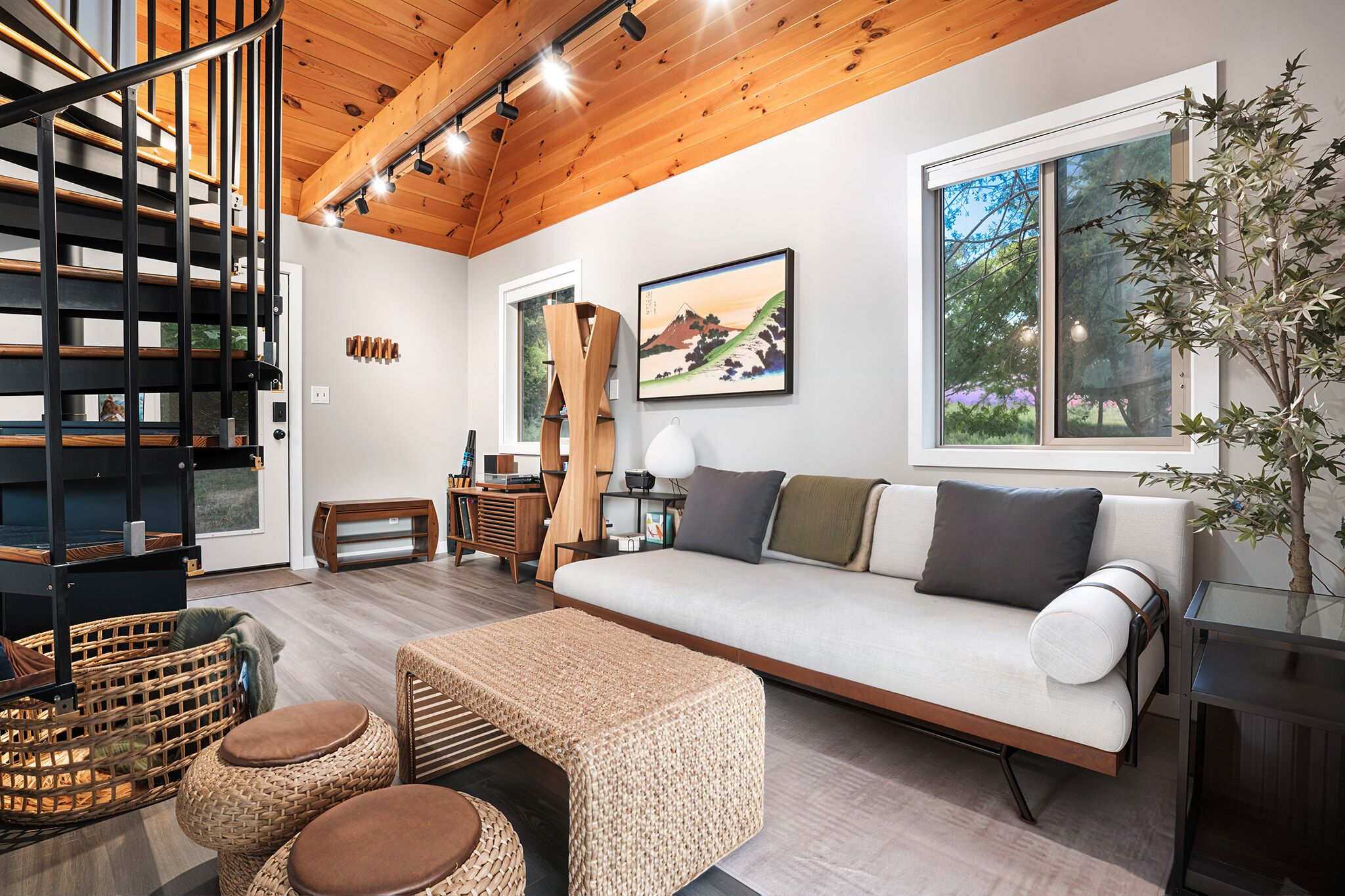 A modern living room with a wooden staircase, a sofa, wicker furniture, and a large window.