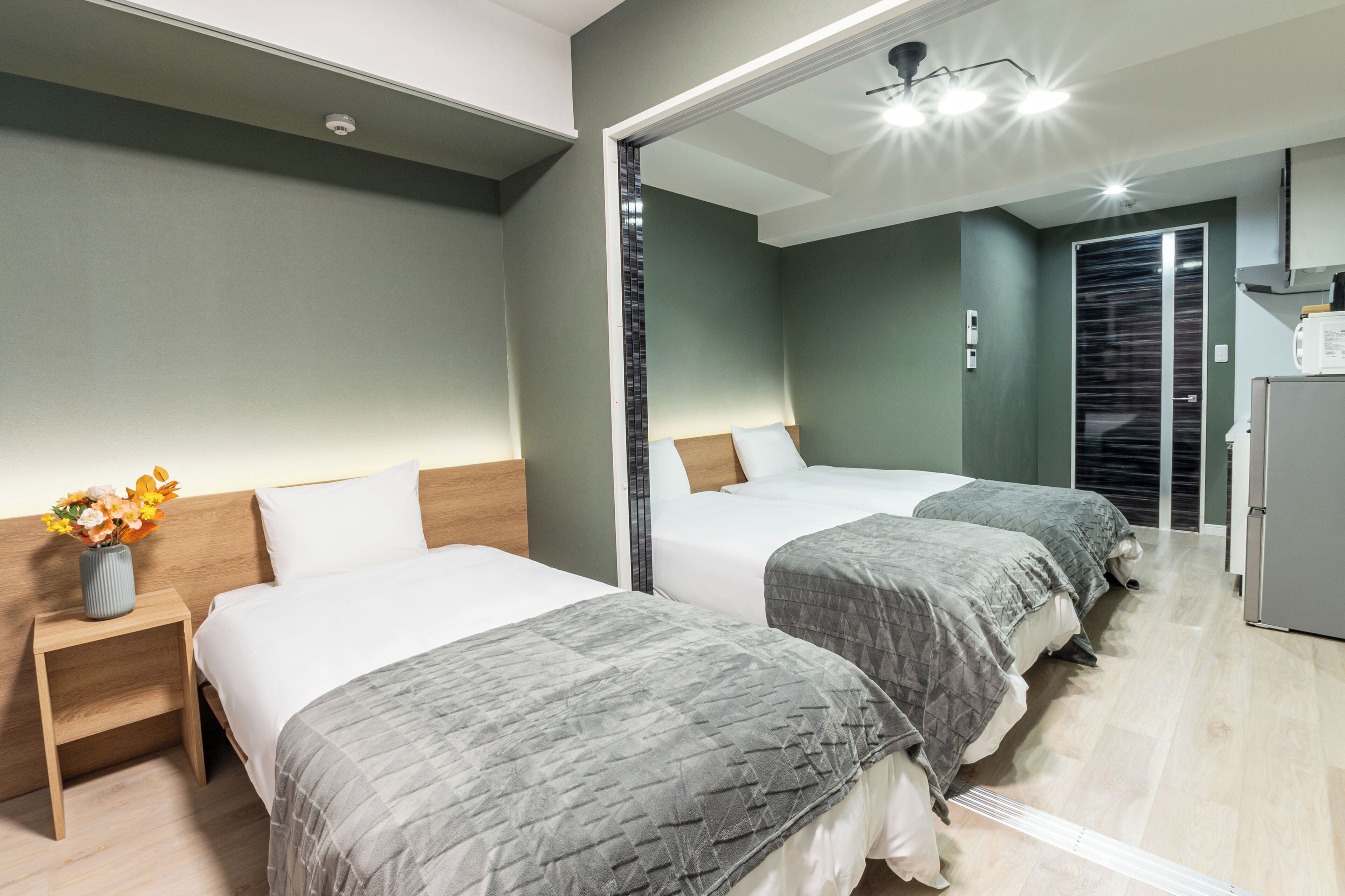 Una habitación de hotel con dos camas, una mesita de noche con un florero, un espejo y una zona de kitchenette.