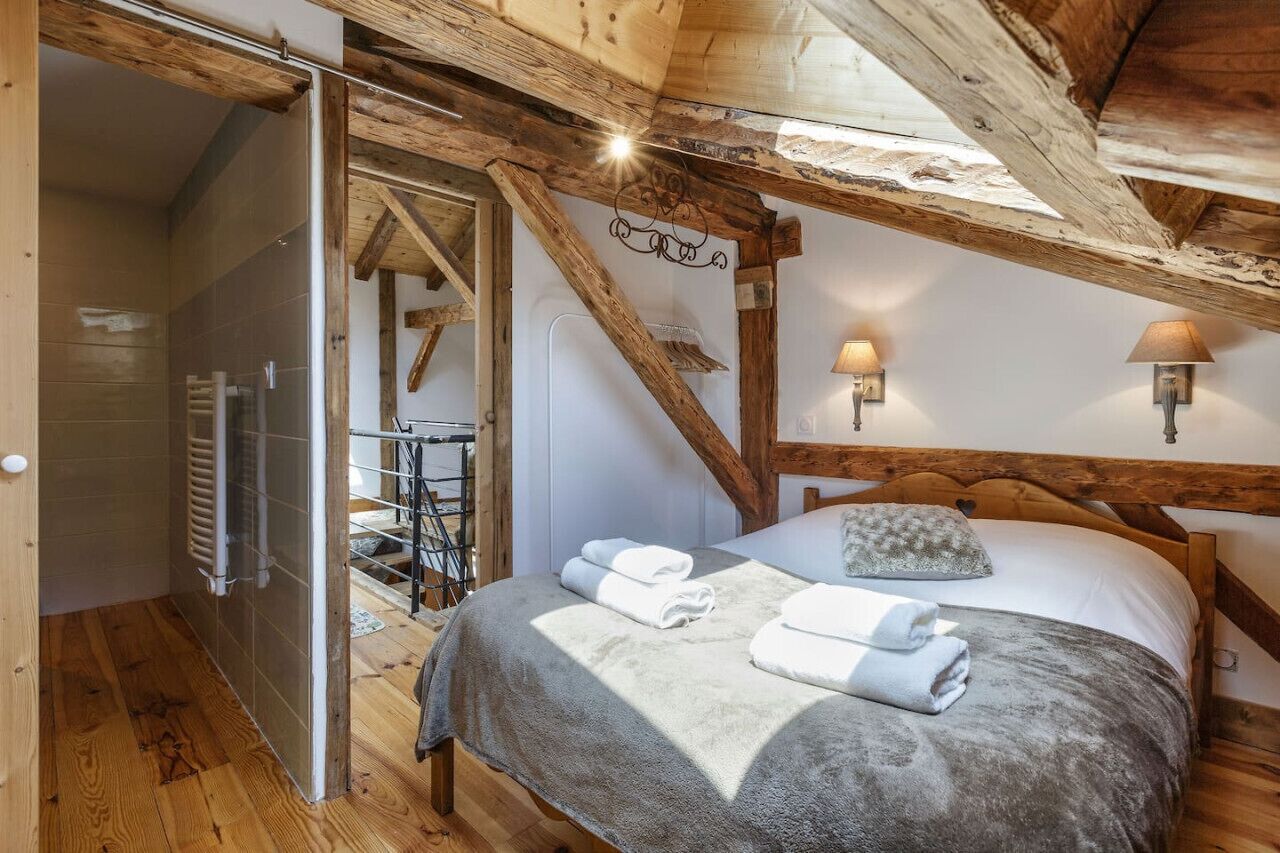 Une chambre avec un plafond en bois, un lit recouvert d’une couverture grise, deux lampes fixées au mur et un porte-serviettes.