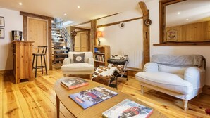 Ein gemütliches Wohnzimmer mit Holzboden, einem weißen Sofa, einem Ledersessel und einem Couchtisch mit Zeitschriften. Eine Treppe mit Holzwand führt in einen anderen Raum.