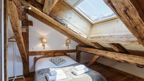 Ein Schlafzimmer mit schrägen Wänden, Holzbalken, einem Oberlicht, einem Bett mit Kissen, einer Nachttischlampe und Handtüchern.