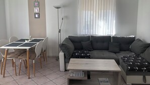 Ett vardagsrum med en soffa, ett soffbord, ett matbord med stolar, en golvlampa och ett fönster med gardiner.