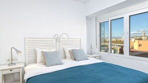 A bedroom with a large bed, white headboard, and a view of a cityscape through large windows. - Solaga - Martinez Virel (M álaga)