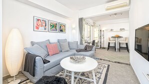 A modern living room with a grey sofa, a white coffee table, and a large floor lamp. There are framed artworks on the wall and a dining area in the background. - Solaga - Martinez Virel (Málaga)