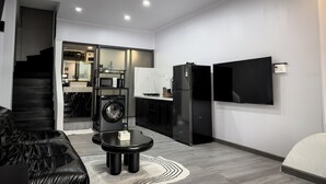 Een knus woongedeelte met een bank, een salontafel, een wasmachine, een koelkast en een aan de muur bevestigde televisie.